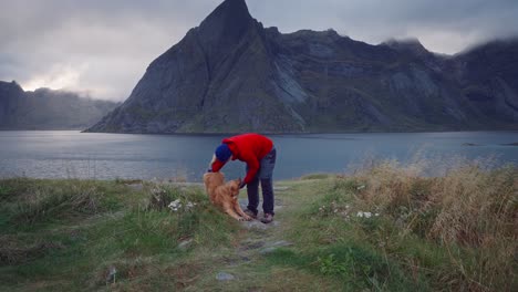 A-young-man-in-a-red-jacket-petting-his-golden-retriever-dog-in-front-of-the-mountains-and-sea-of-Hamnøy-near-Reine,-Lofoten-Islands,-Norway