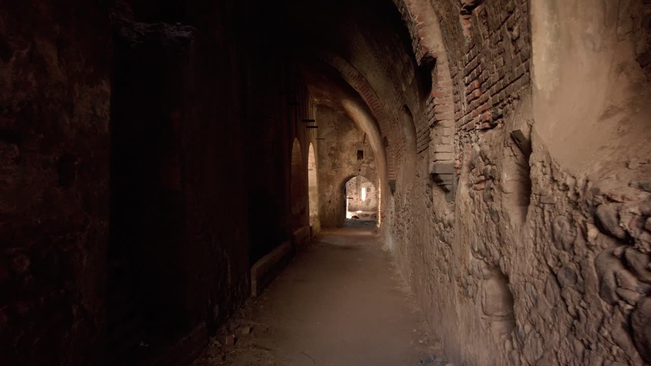 Vista interior de um corredor de fazenda abandonada com arcos repetidos e paredes de pedra texturizadas, que conduz a uma passagem estreita com luz natural e uma atmosfera rústica envelhecida.