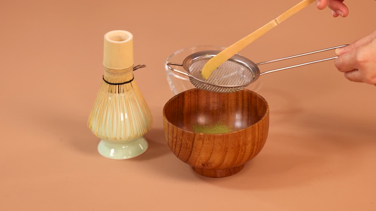 Hand sifts matcha tea powder into wooden bowl using bamboo scoop, minimal style, soft lighting