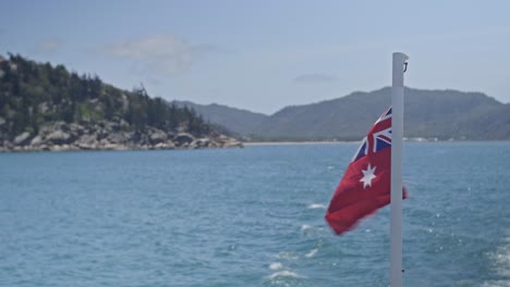 a red flag on a flagpole waving in the breeze on the back of a ferry leaving an island