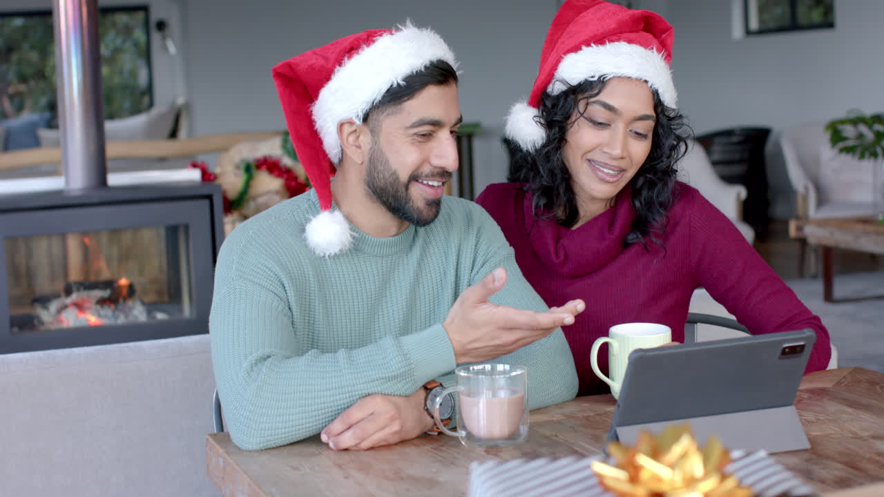 una feliz pareja biracial con sombreros de papá noel usando una tableta para una llamada de video en casa, en cámara lenta