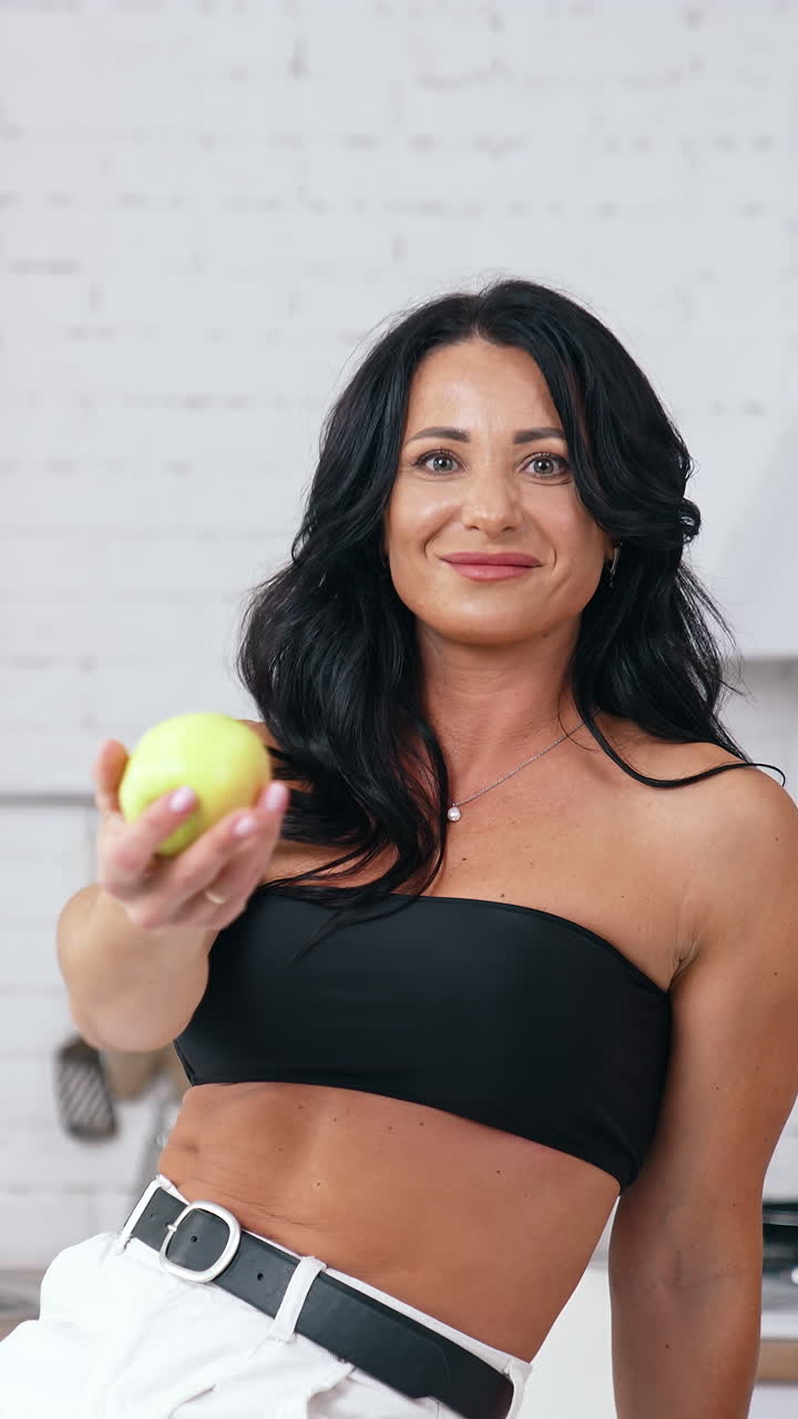 Gorgeous brunette woman sitting at the edge of the table and looking at the healthy apple. Diet and vegan food concept Vertical video