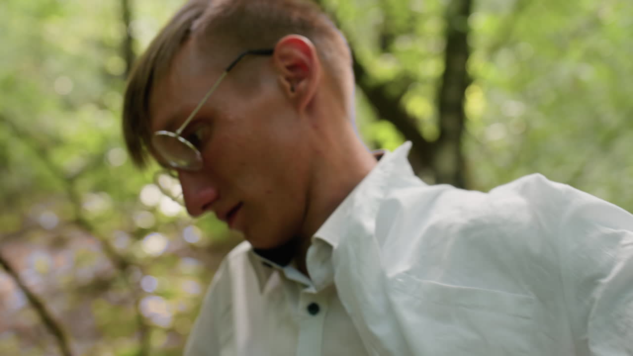Closeup view of young man in white shirt wearing coat in forest surrounded by lush greenery and soft blurred background, emphasizing natural environment, outdoor lifestyle