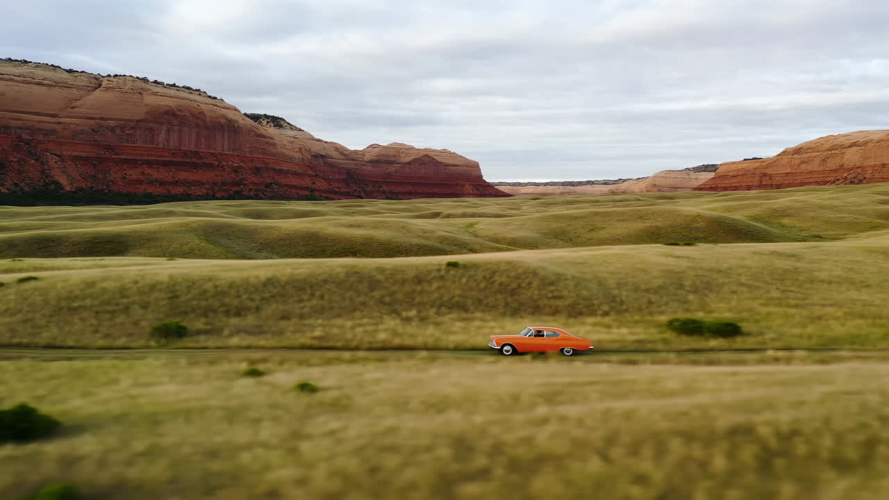 Orangefarbenes Auto durchquert weites Graslandtal mit roten Felsklippen unter stimmungsvollem bedecktem Himmel, kinoreife Seitenansicht