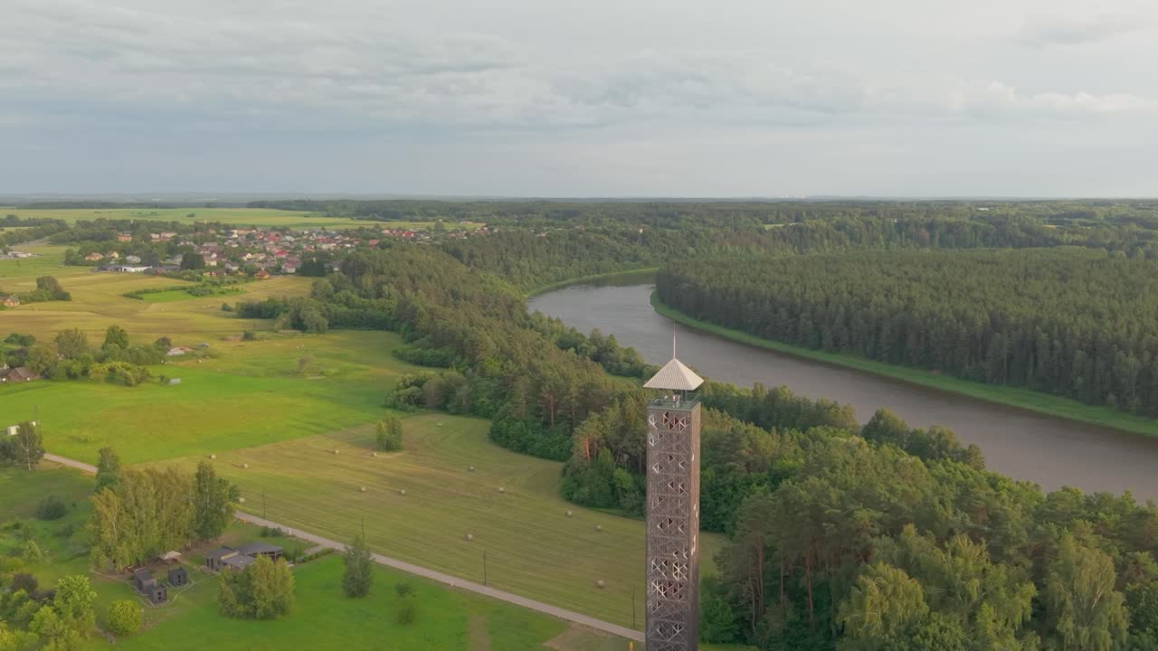 Aerial view of wooden observation tower, river bend, lush green forest and village under cloudy summer sky in Birstonas Lithuania, orbit shot