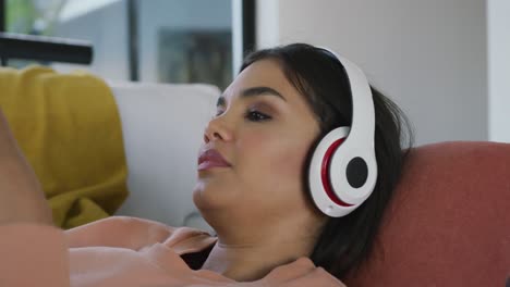 Video-of-relaxed-biracial-woman-lying-on-sofa-with-headphones-and-listening-to-music