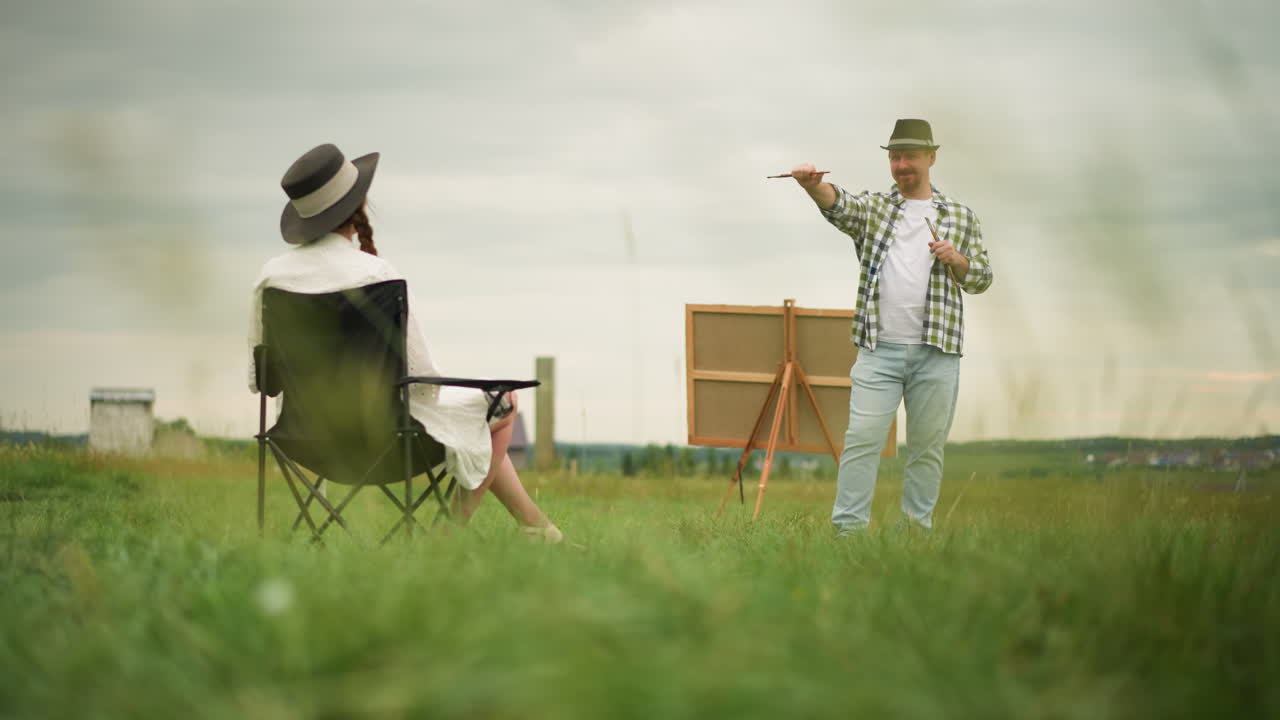 체크 된 셔츠를 입고 브러쉬를 들고 있는 공예인 은 잔디 에 있는 의자 에 앉아 있는  옷 과 모자 를 입은 여자 를 향해 브러쉬 를 가리키면서 미소 짓는다