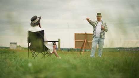 a craftsperson wearing a checked shirt and holding a brush smiles while pointing the brush at a woman dressed in a white gown and hat, sitting on a chair in a grassy field