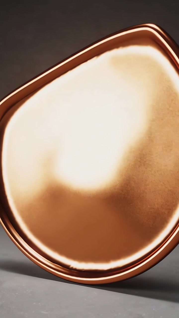 Video verticale: Sequenza iniziale di una goccia di bronzo che si inclina e ruota su una piattaforma grigia rivelando il bordo