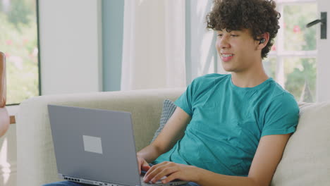 teenage boy wearing wireless earbuds making video call on sofa at home using laptop