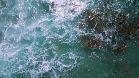 Vista-Aérea-Desde-Arriba-Del-Dron-Que-Muestra-Las-Olas-Formando-Espuma-Y-Rompiéndose-Sobre-Un-Afloramiento-Rocoso-Poco-Profundo-En-El-Agua