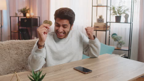 un joven indio celebra la victoria de la criptomoneda mostrando bitcoin después de comprobar los gráficos de intercambio