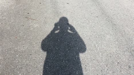 man films his shadow walking along an asphalt road with his phone