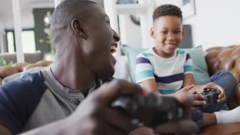 Video-of-happy-african-american-father-and-son-sitting-on-sofa-and-playing-video-games