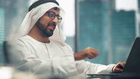 successful arab businessman in glasses and white traditional outfit sitting in office and working on laptop. business manager planning corporate strategy. saudi, emirati, arab businessman concept.