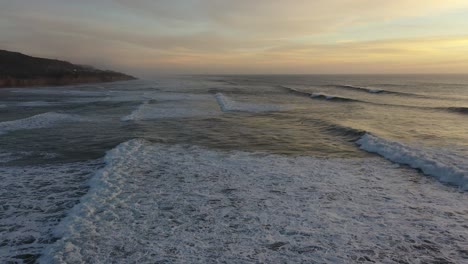 Dramatic-drone-shot-flying-just-above-crashing-waves-during-sunset,-slow-pan-up-reveals-the-beauty-of-the-seascape-and-the-warm-colors-of-the-setting-sun