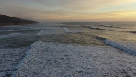 dramatic drone shot flying just above crashing waves during sunset, slow pan up reveals the beauty of the seascape and the warm colors of the setting sun