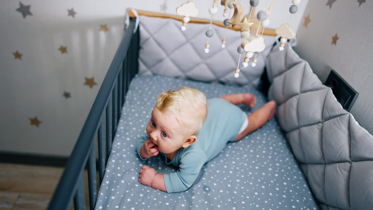 Tiny infant baby lies in the crib chewing his little fist. Beautiful male child in a cozy cot with mobile. Top view.