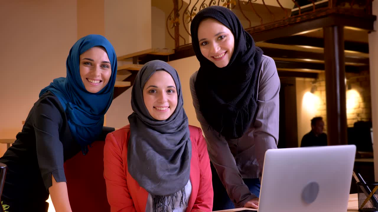 히자브를 입은 세 명의 젊은 이슬람 여성 사무실 직원들이 직선적으로 카메라를 바라보고 웃으며 작업장에서 노트북 앞에 있는 클로즈업 초상화