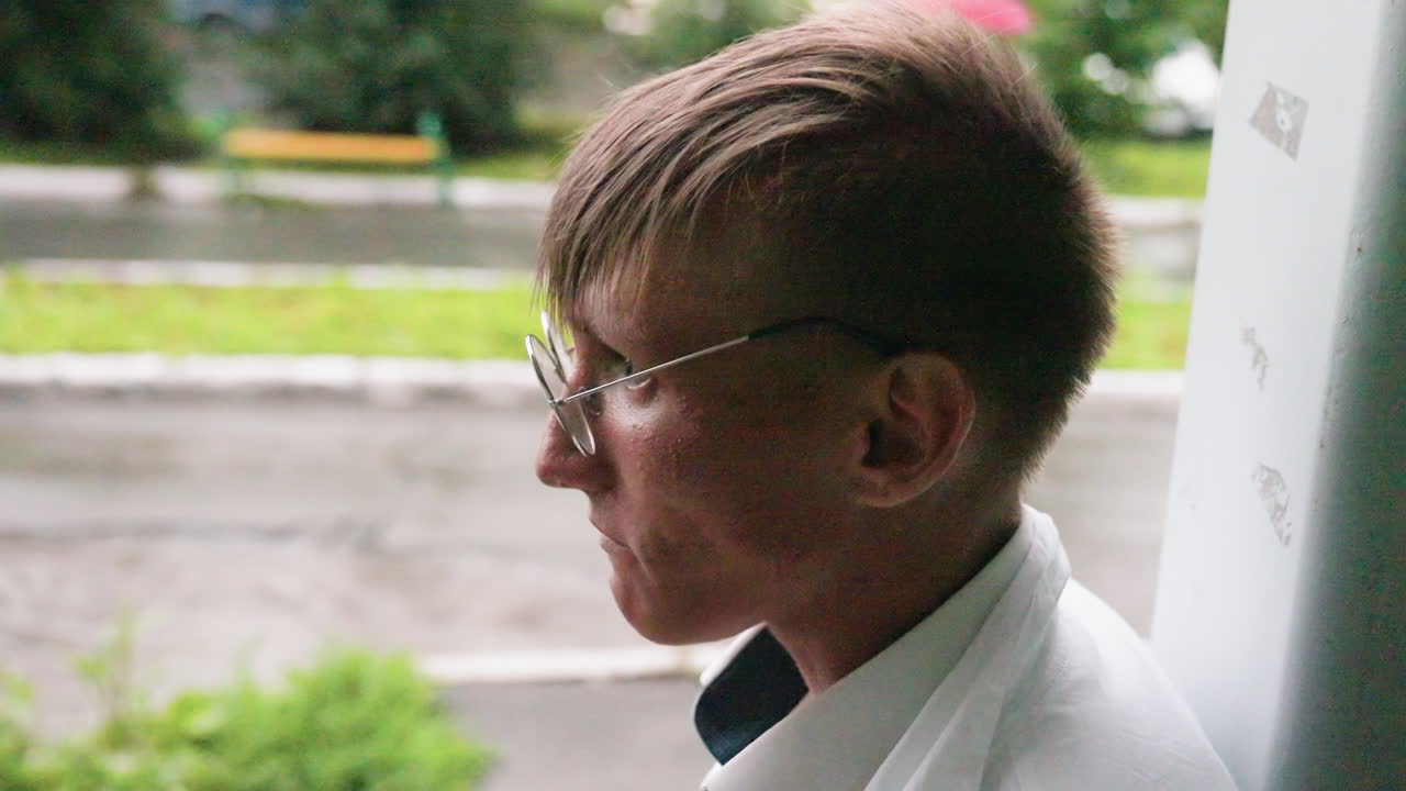 Young botany student in white coat and glasses drinks unusual green beverage from bottle during outdoor break, enjoying refreshing hydration in relaxed urban natural environment near greenery