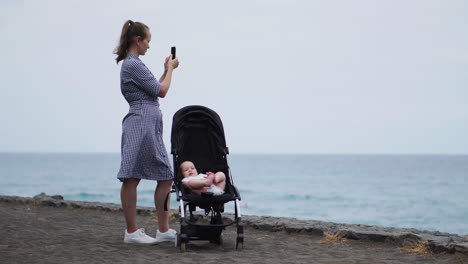 a young mother stands with a baby sitting in a wheelchair near the ocean and takes pictures on a smartphone beautiful photos for social networks and blog