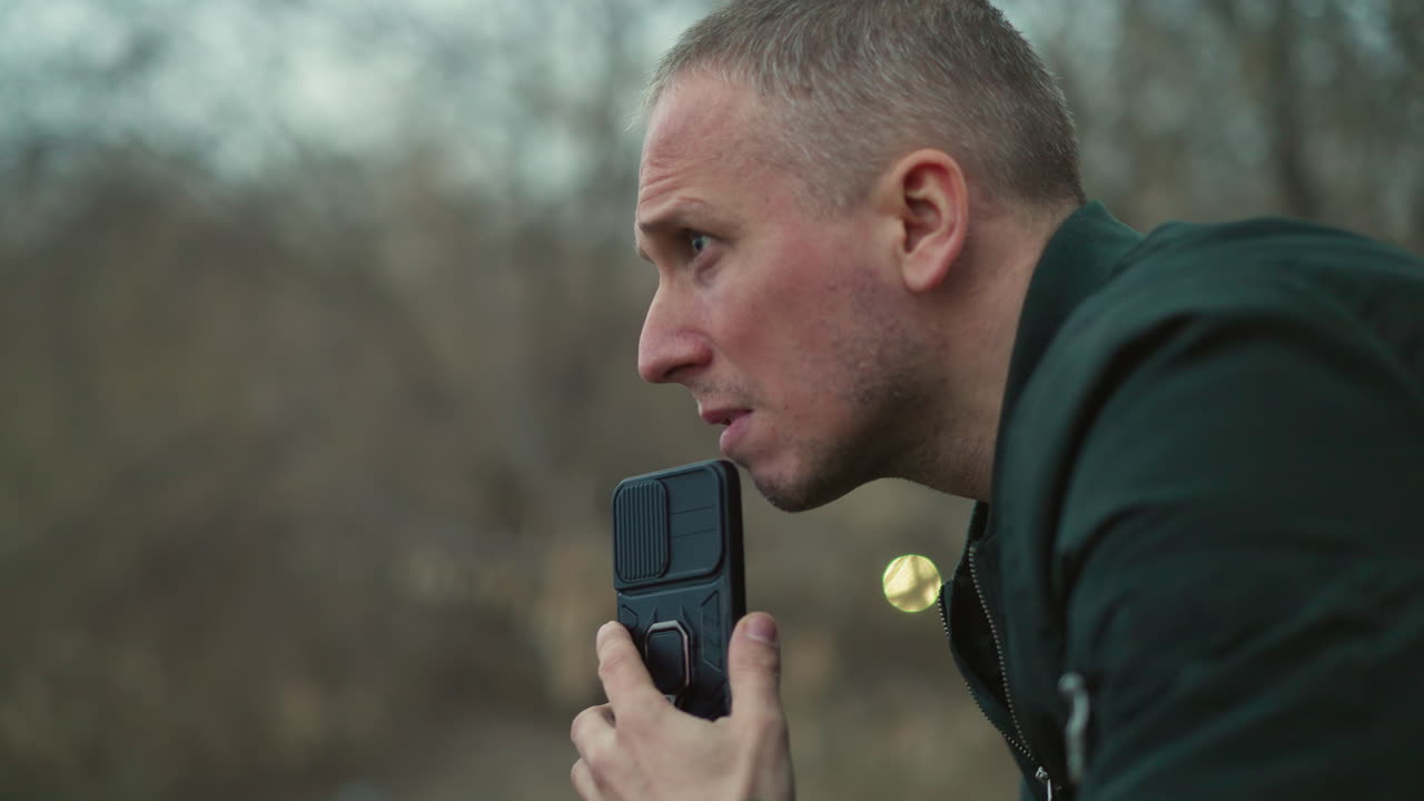 A close view of a man looking intensely to the side, holding a black smartphone near his mouth, the background features a forest, with two blur ball like yellow light seen