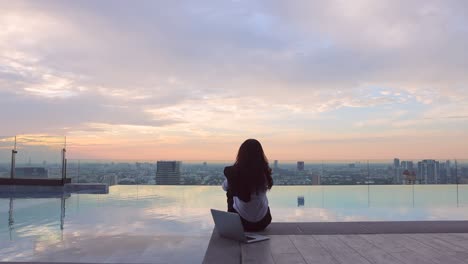 businesswoman dressed in formal clothes with suit over her shoulder sitting on the edge dangling her feet in swimming pool on skyscraper. woman using social media on laptop for relax during sunset.