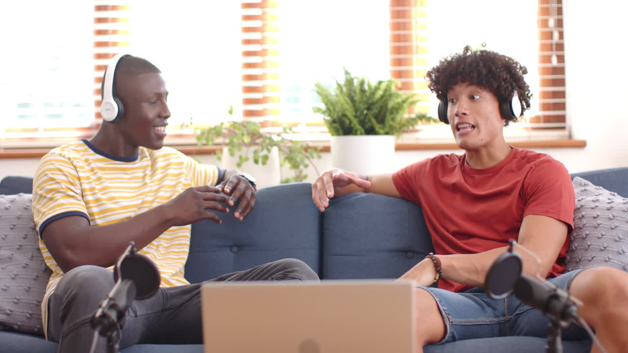 Listening to music, two multiracial male friends relaxing with headphones and laptop, at home