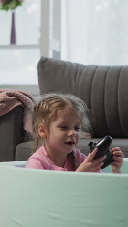 boy and girl throw plastic balls actively sitting in dry pool in light living room. satisfied children siblings find joystick and examine device with interest