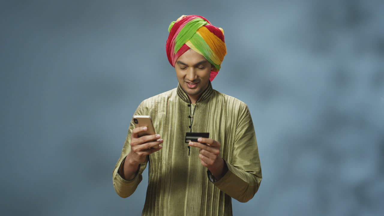apuesto hombre indio alegre en turbante y ropa tradicional comprando en línea con smartphone y sonriendo a la cámara