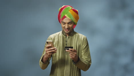 apuesto hombre indio alegre en turbante y ropa tradicional comprando en línea con smartphone y sonriendo a la cámara