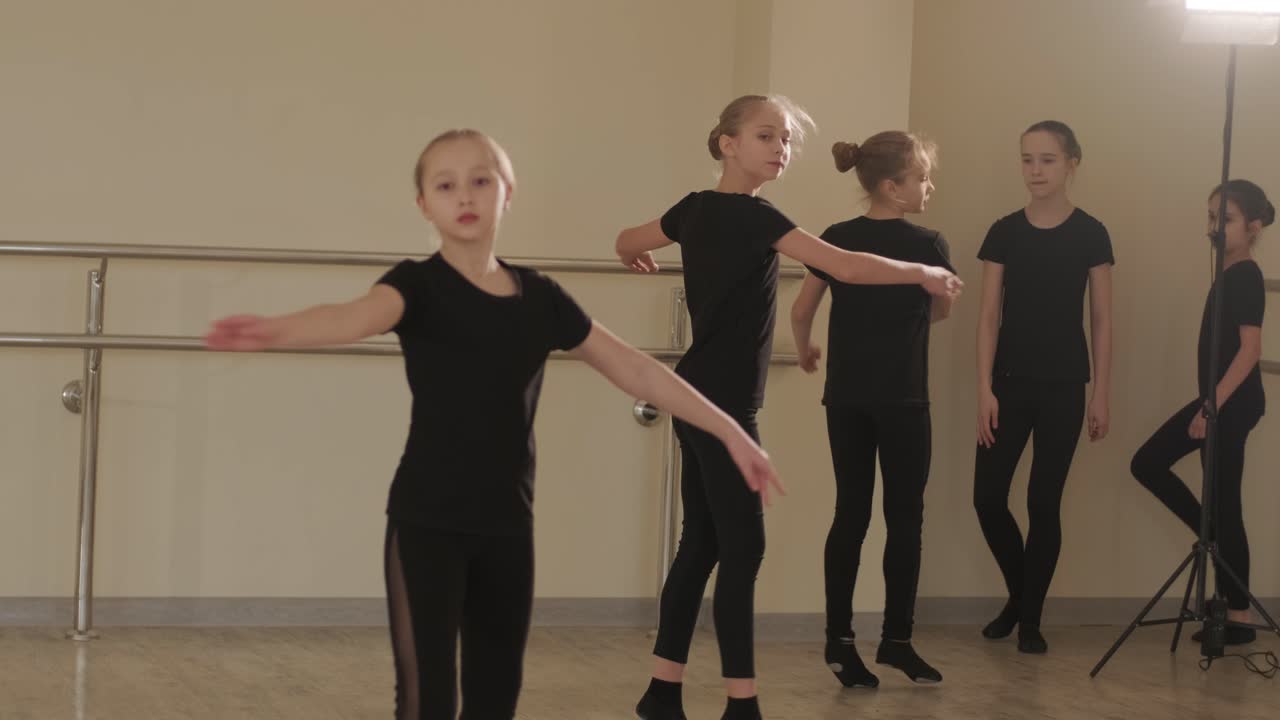 A group of young ballet students in black dancewear practicing positions in a spacious ballet studio with wooden flooring and wall-mounted barres. Focused expressions and synchronized movements.