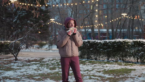 young girl standing in park holding napkin while eating corn, outdoor winter scene with snow on ground, warm light bokeh in background