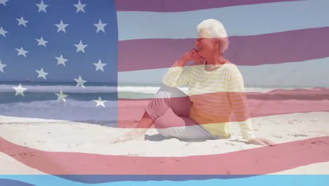 Animation-of-smiling-senior-caucasian-woman-sitting-on-beach-over-flag-of-united-states-of-america