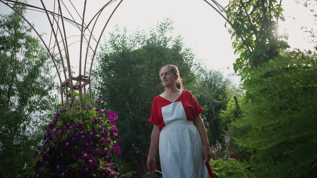 Kaukasisk kvinde i hvidt forklæde læner sig op ad en jernspalier, omgivet af rigelige lilla blomster og frodige buske. Solbelyst modlys, et mildt smil, bærer en kurv med blomster, havefestival.