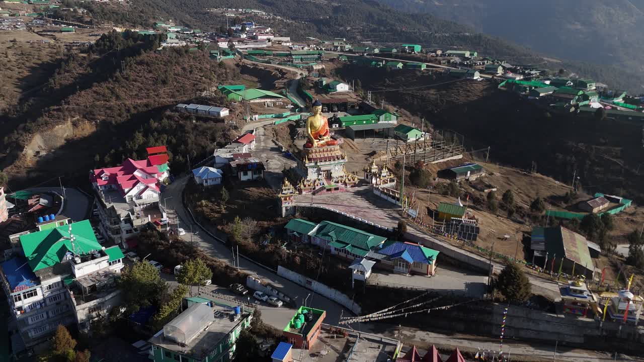 Drone görüntüleri, Himalaya dağlarında kıvrımlı yollar ve Budist manastırı yapılarıyla iç içe geçmiş, renkli yeşil ve kırmızı çatılı tepelik bir kasabanın üzerinde yükselen büyük bir altın Buda heykelini ortaya koyuyor.