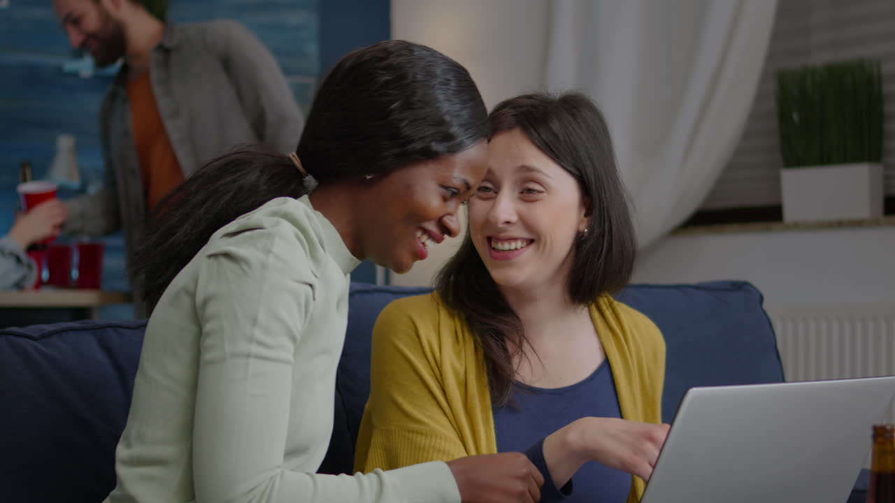 Two multiethnic women sitting on couch watching comedy series on laptop