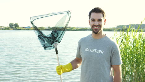 Joven-Apuesto-Sonriendo-Y-Posando-Para-La-Cámara-Con-Una-Red-De-Acero-Llena-De-Basura-Después-De-Limpiar-El-Fondo-Del-Lago