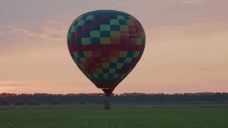 wide farmland landscape at dusk showing colorful hot air balloon hovering low with flame flicker beneath envelope against soft pastel sky creating tranquil rural adventure