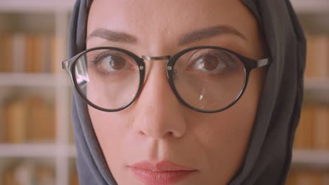 closeup portrait of young arabian female face in glasses and hijab looking at camera in library indoors