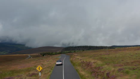 Forwards-tracking-of-vintage-car-driving-on-road-surrounded-by-moorlands.-Highlands-landscape-with-hills-shrouded-in-clouds.-Ireland