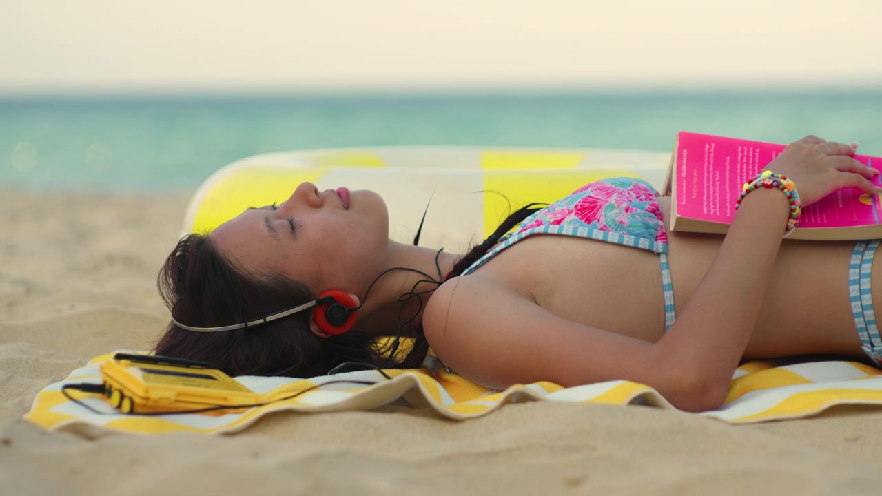 Young Asian woman in colorful bikini relaxing on beach towel with headphones listening to music relaxing from reading book beside turquoise sea on a sunny day, static sideview