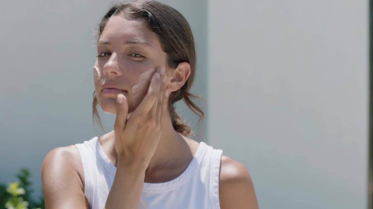 Raising right hand woman smoothing pale cream with applicator on pale wall, wearing white tank top