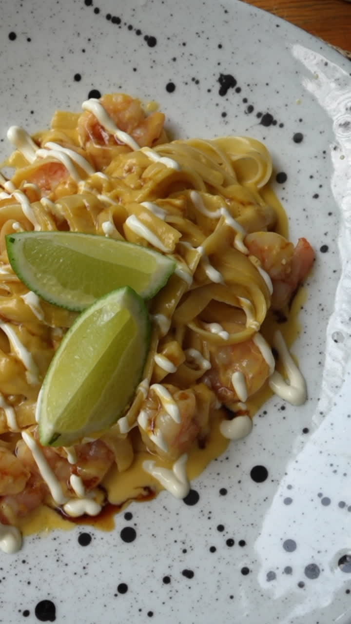 Vertical video capturing a close-up shot of flavorful Colombian shrimp pasta garnished with lime and drizzled sauce. Bright lighting enhances the delectable presentation
