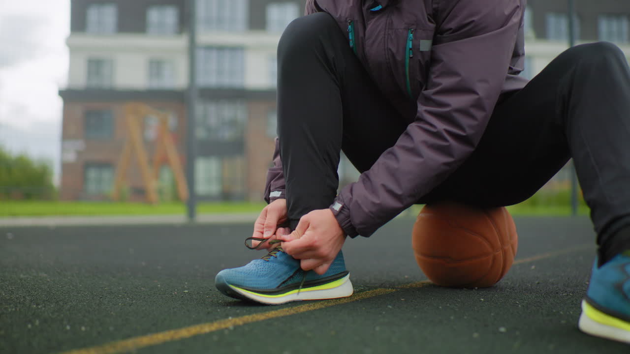 Corridore che si allaccia la scarpa su un campo all'aperto, seduto su un pallone da basket, che indossa un berretto e una giacca, scarpe da ginnastica blu sull'asfalto bagnato, palazzi sullo sfondo, routine pre-corsa concentrata mentre sistema i lacci preparandosi