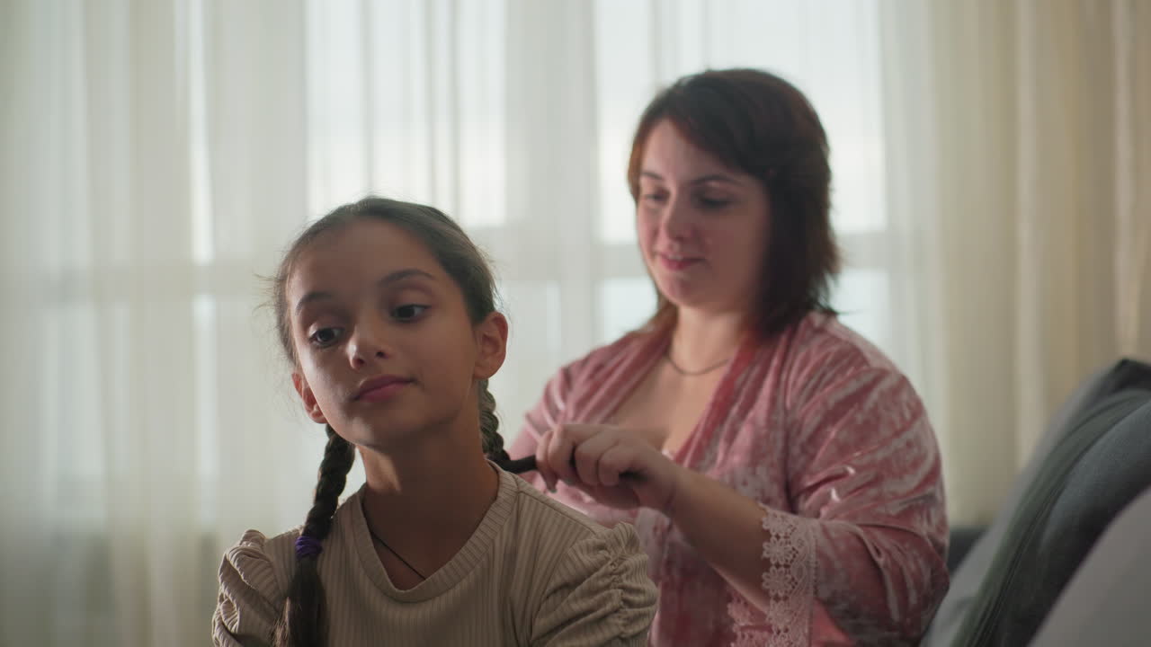 mum braiding daughter’s hair on sofa close-up gentle grooming moment in sunlit living room portraying tender mother child bond warm home atmosphere calm intimate personal care scene