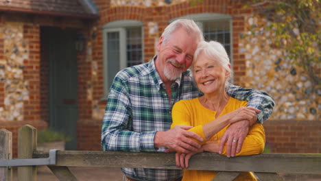 Portrait-Of-Happy-Retired-Senior-Couple-Outside-Home-Leaning-On-Gate