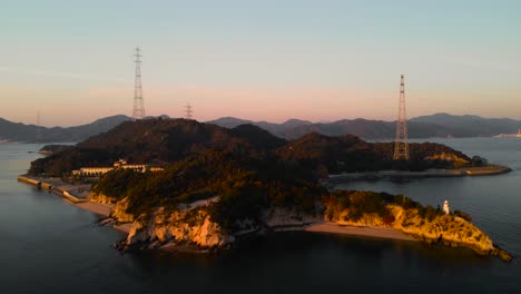 Rückwärtsbewegung-Der-Luftdrohne-über-Der-Insel-Mit-Leuchtturm-Und-Stromleitungen-Bei-Sonnenuntergang