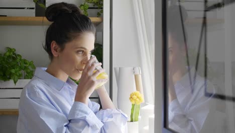 mujer joven mirando por la ventana y bebiendo jugo de naranja en casa