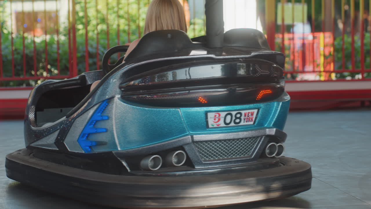 若いドライバーがティール色のバンパーカーを運転している。赤い光漏れとモーションブラーがかかり、家族が応援する活気ある公園の週末の風景。ハンドル操作とスピードを示すクローズアップの後方アングルで、臨場感あふれる高速アトラクションの様子。